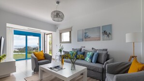 Ein modernes Wohnzimmer mit einem weißen Couchtisch, grauen Sofas mit gelben Kissen, einem großen Fenster mit blauem Rahmen und Blick aufs Meer.