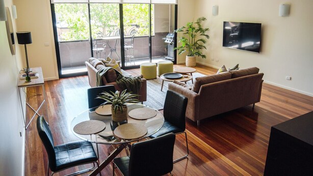 A modern living room with a glass dining table, two sofas, a TV, and a sliding glass door to the outside. - Spacious, modern apartment right in the Bendigo CBD (Bendigo)