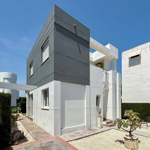 Ein modernes, zweistöckiges Haus mit flachem Dach, einem Balkon und einer kleinen Gartenanlage mit Topfpflanzen.