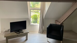 A room with a TV on a white table, a black chair, and a window with a view of greenery. - Chambre Bonheur (Romeries)