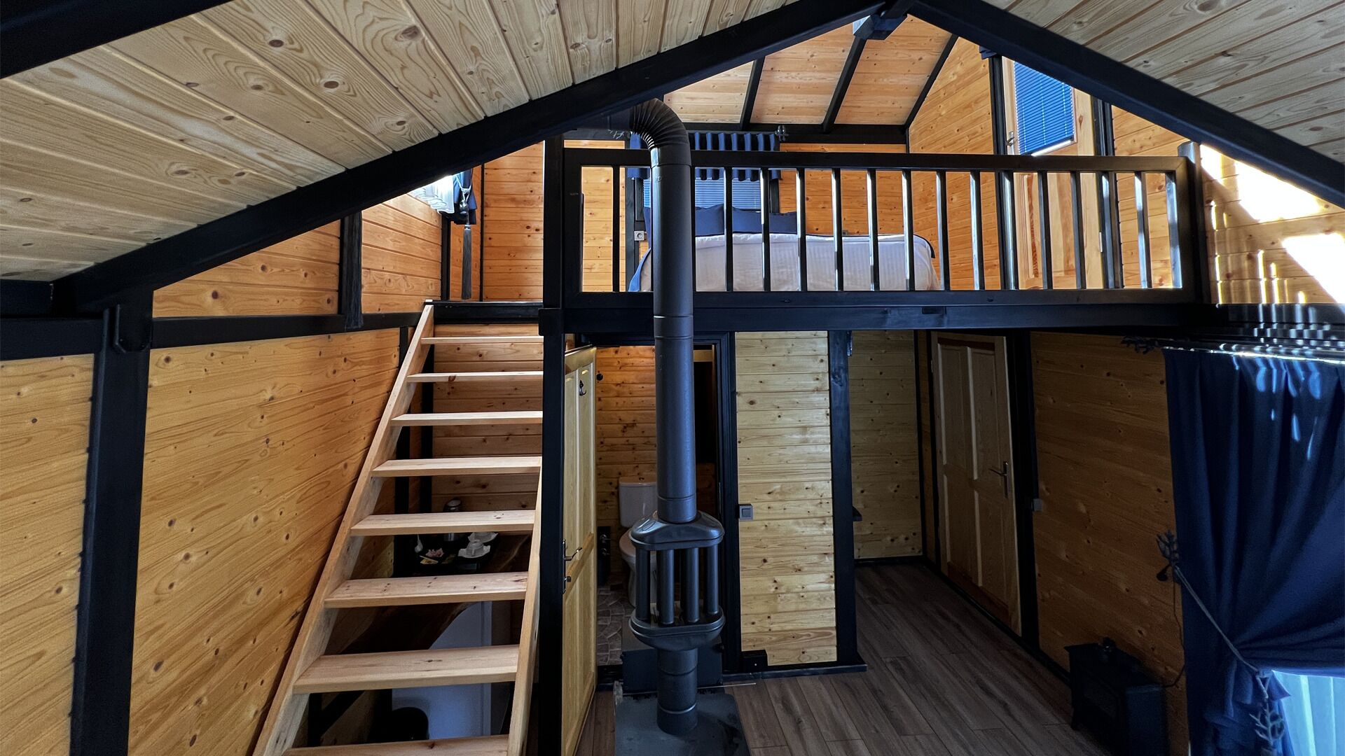 Une cabane en bois avec une chambre à coucher située en mezzanine, un escalier et un poêle à bois.