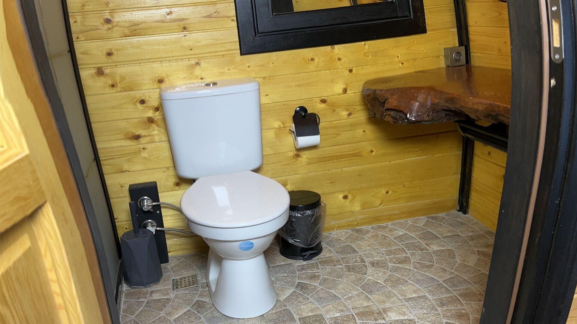 Un baño pequeño con inodoro blanco, un cubo de basura y un dispensador de jabón en una pared de madera.
