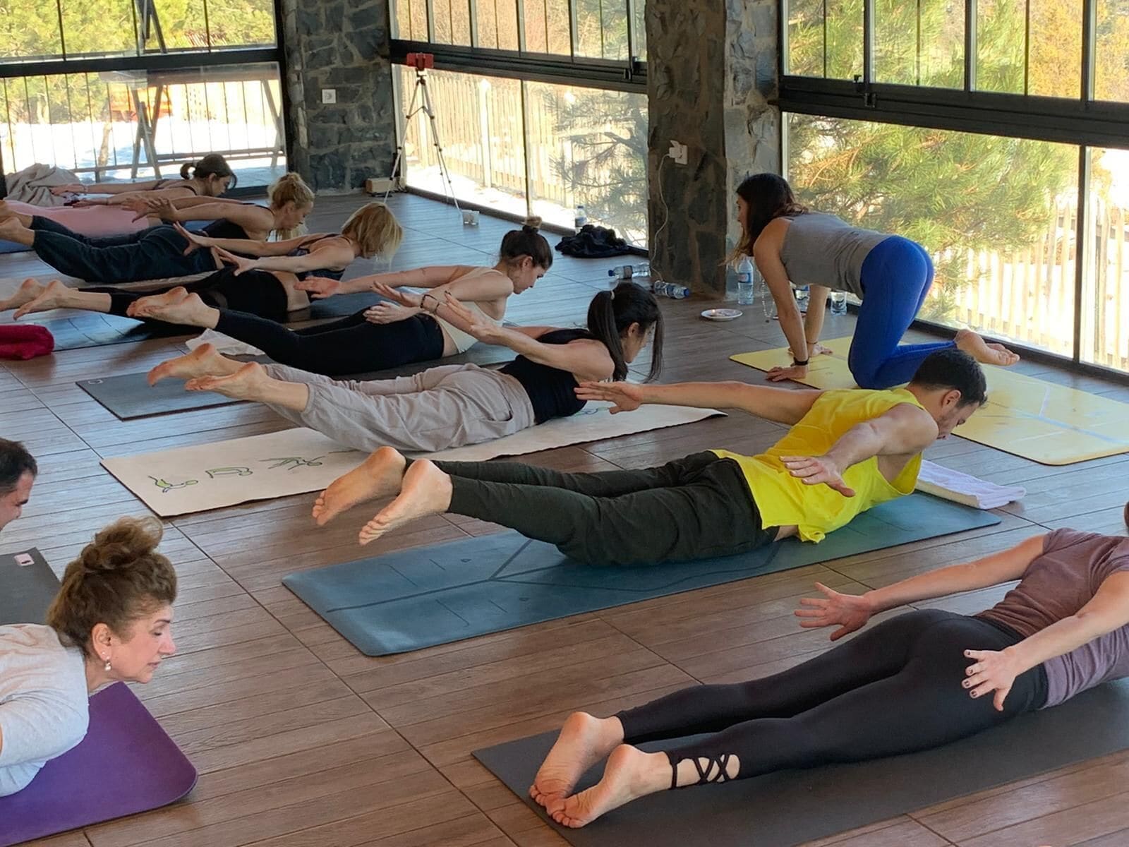 Um grupo de pessoas em uma aula de yoga sobre tapetes em um espaço com janelas amplas e piso de madeira.
