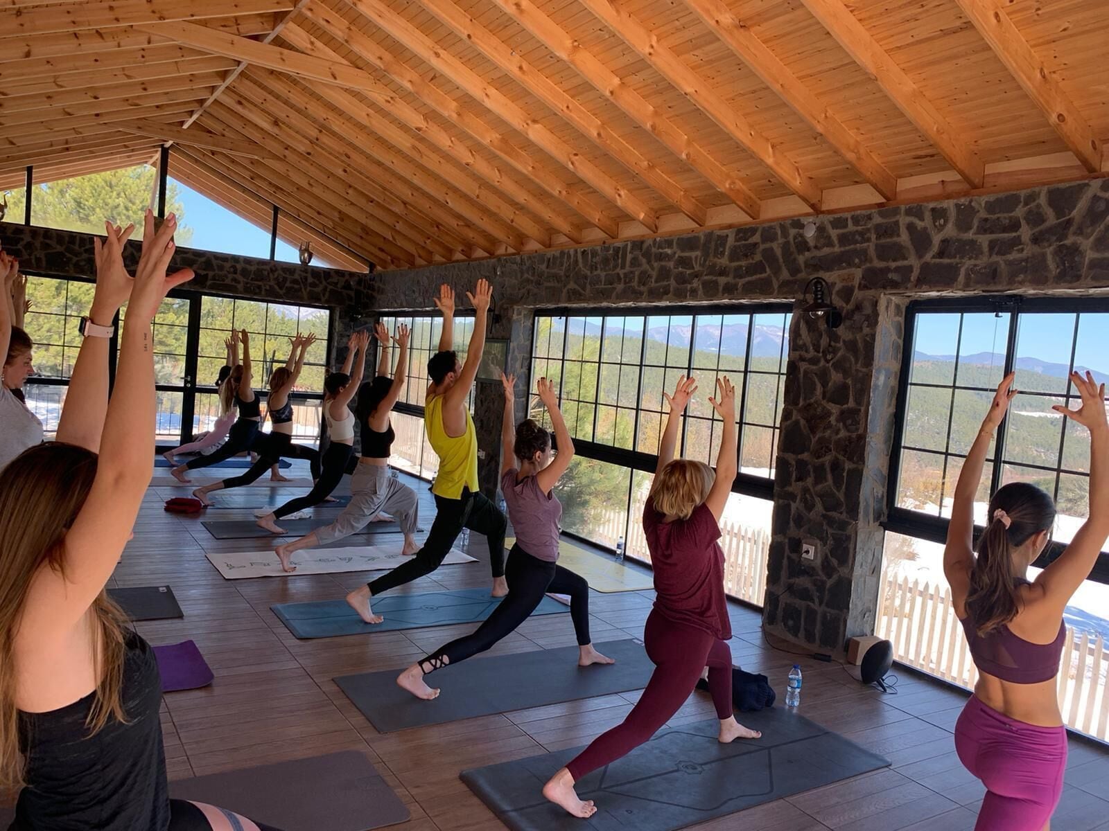Aula de yoga em um espaço com paredes de pedra, grandes janelas, vigas de madeira no teto e vista para montanhas.