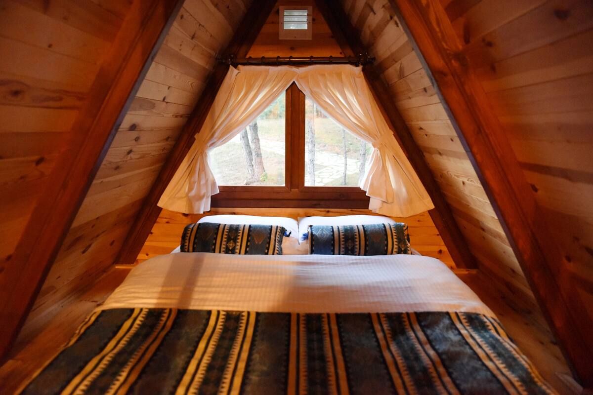 Quarto de cabana de madeira aconchegante, com cama, cortinas e janela com vista para uma floresta.
