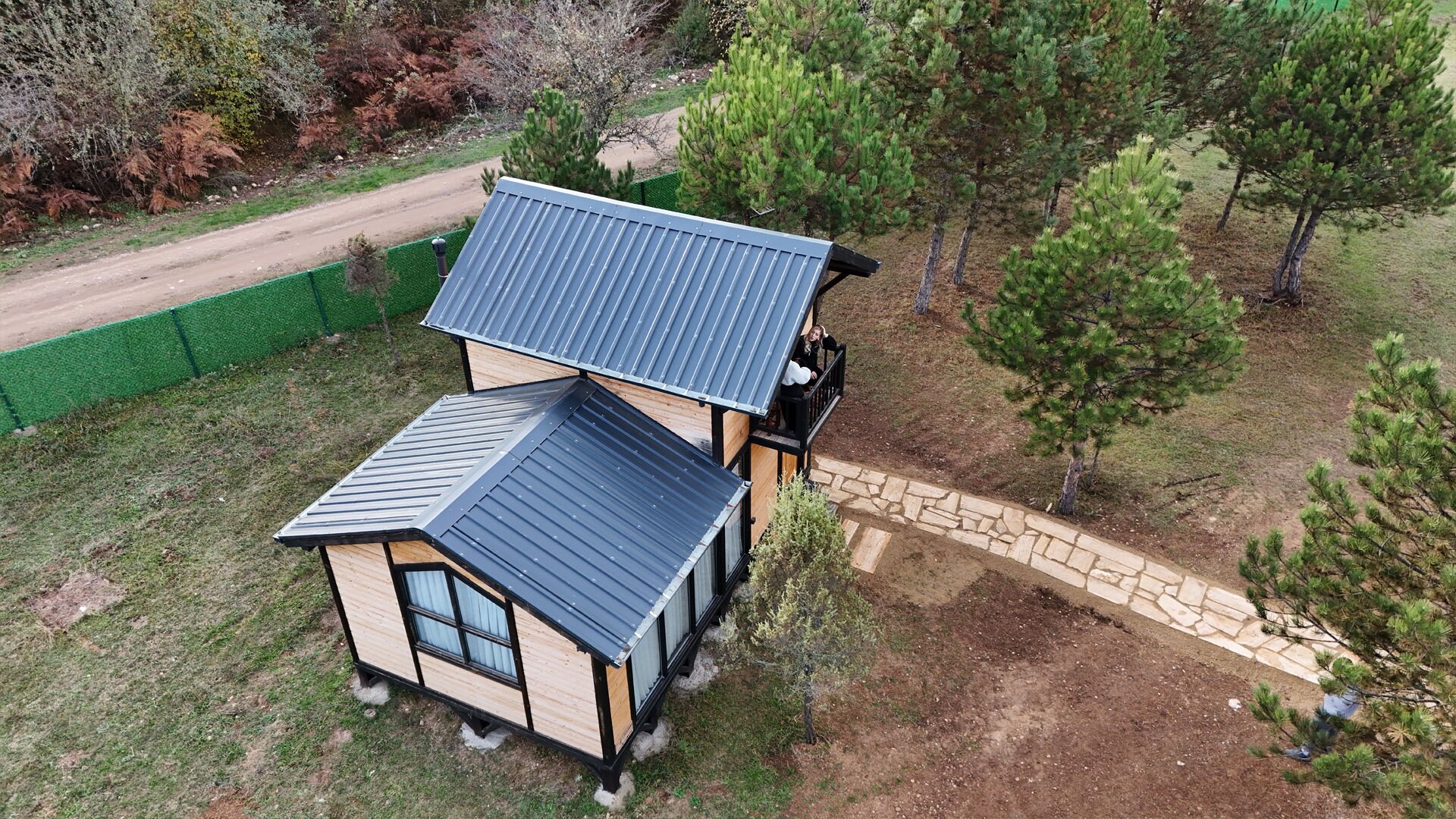 A two-story house with a sloped roof, a stone pathway leading to the entrance, and a fenced yard with pine trees.