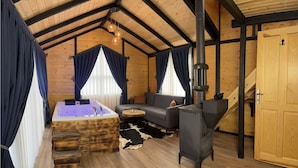 A cozy cabin interior with a wooden ceiling, a large bathtub, a sofa, a small round table, a wood-burning stove, and a staircase.