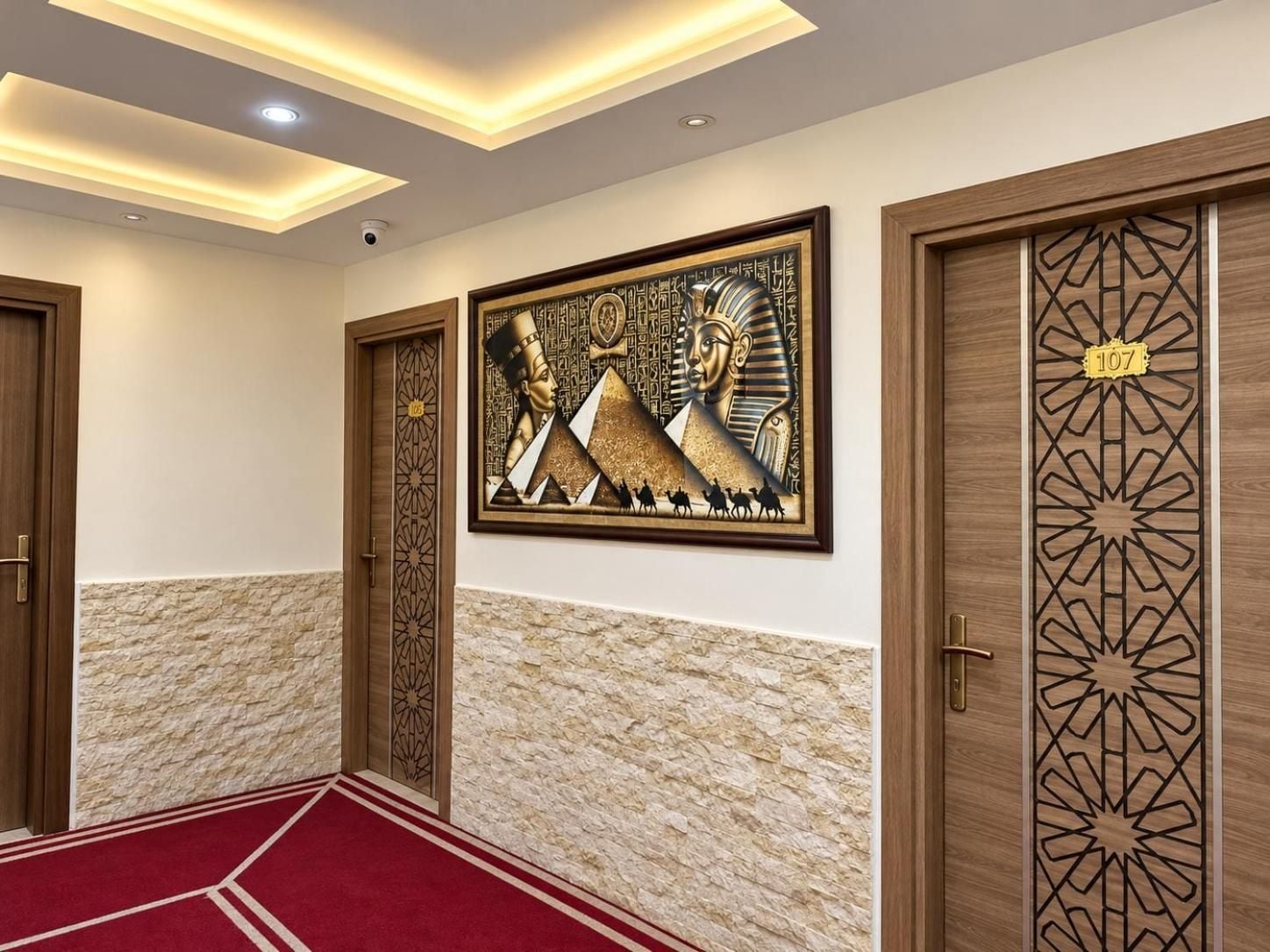 A hallway with a red carpet, wooden doors with intricate designs, and a wall-mounted artwork depicting Egyptian hieroglyphs and pyramids.