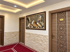 A hallway with a red carpet, wooden doors with intricate designs, and a wall-mounted artwork depicting Egyptian hieroglyphs and pyramids. - Pyramids Gardens Hotel (Giza)