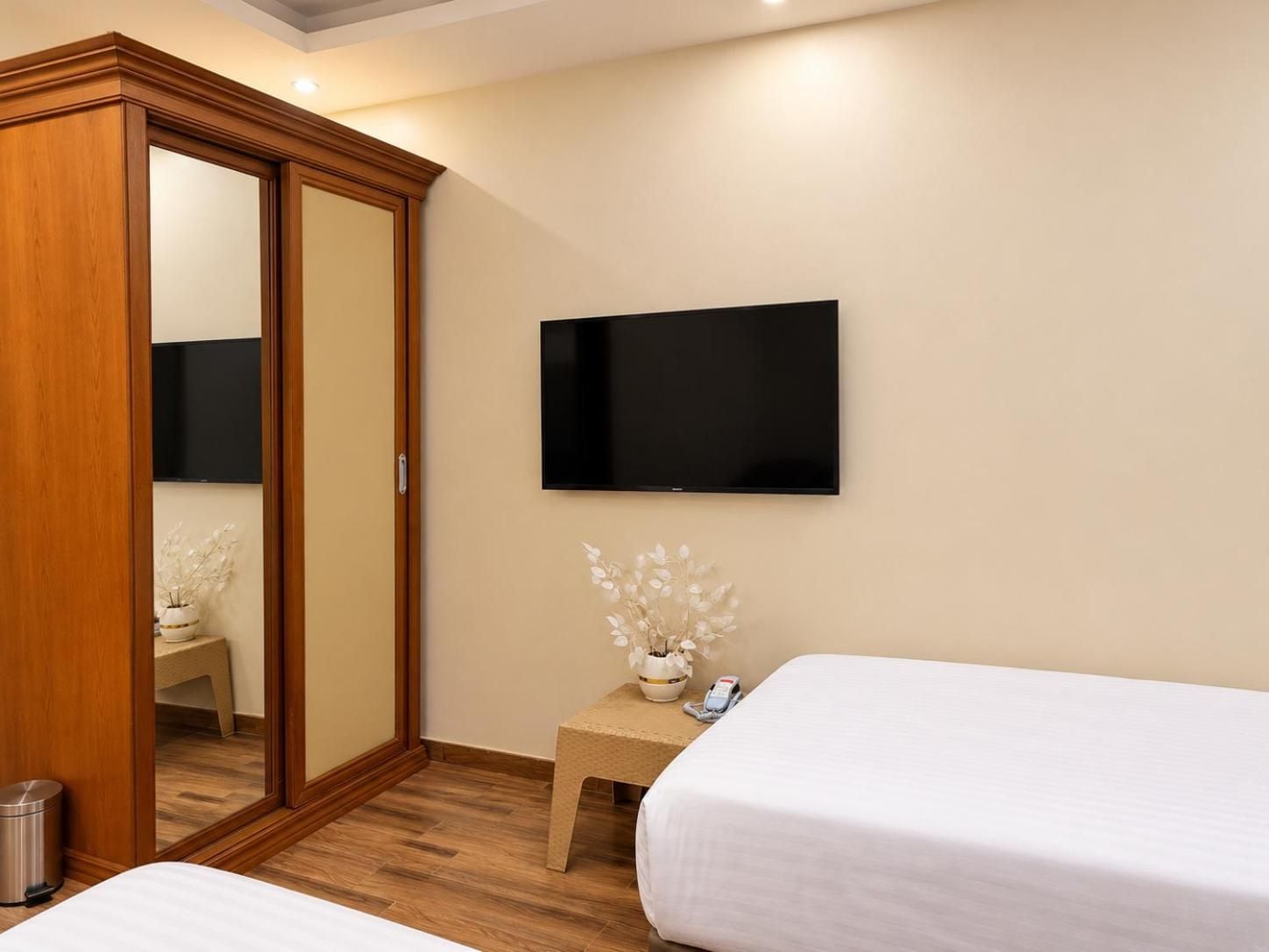 A hotel room with a wooden wardrobe, a flat-screen TV mounted on the wall, a small bedside table with a vase of flowers, and two beds.