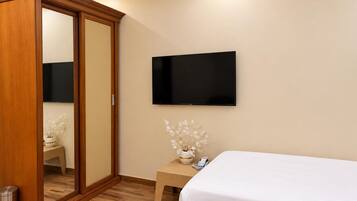 A hotel room with a wooden wardrobe, a flat-screen TV mounted on the wall, a small bedside table with a vase of flowers, and two beds.