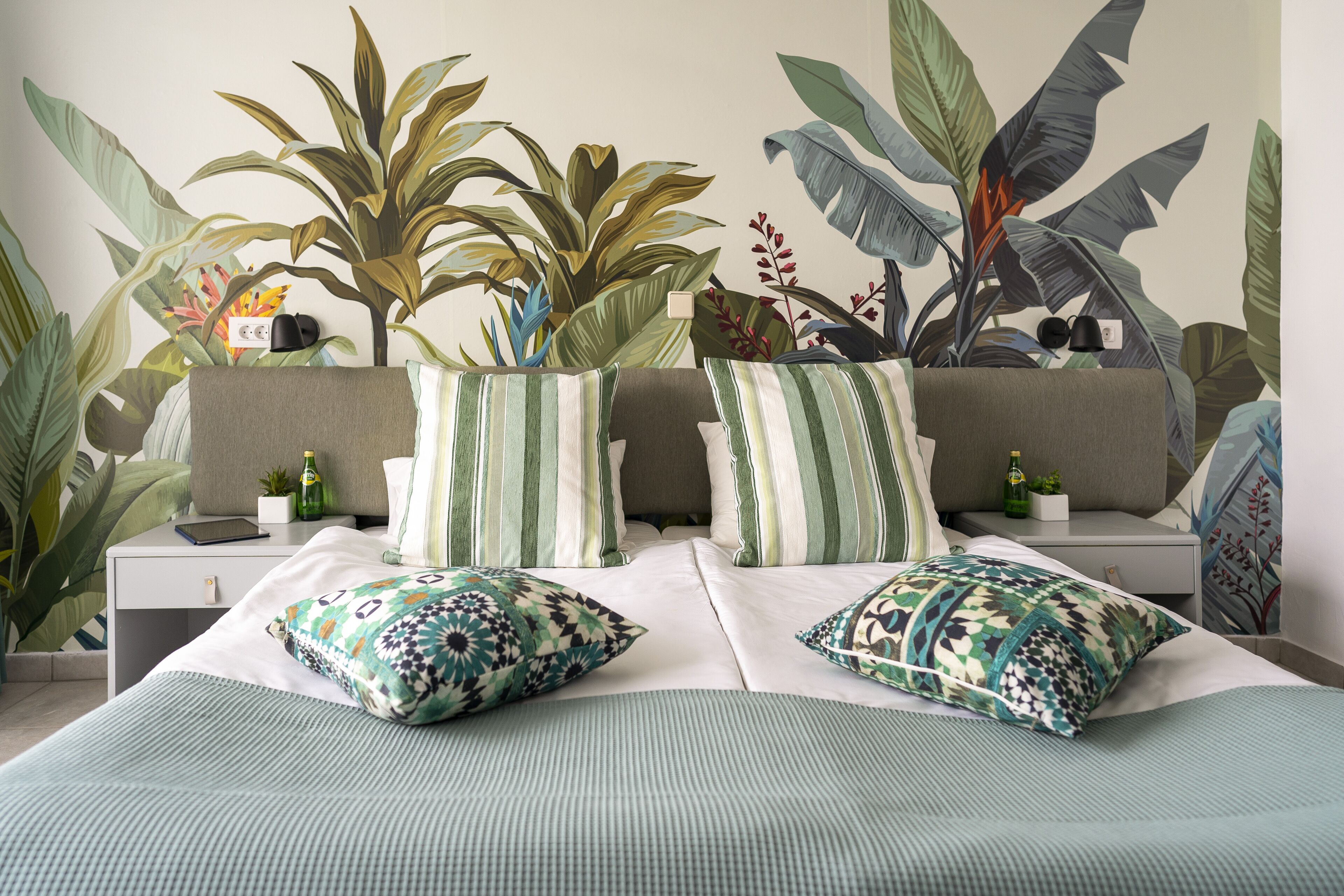 Un dormitorio con una cama, dos mesitas de noche, un sofá y un amplio mural vegetal en la pared.