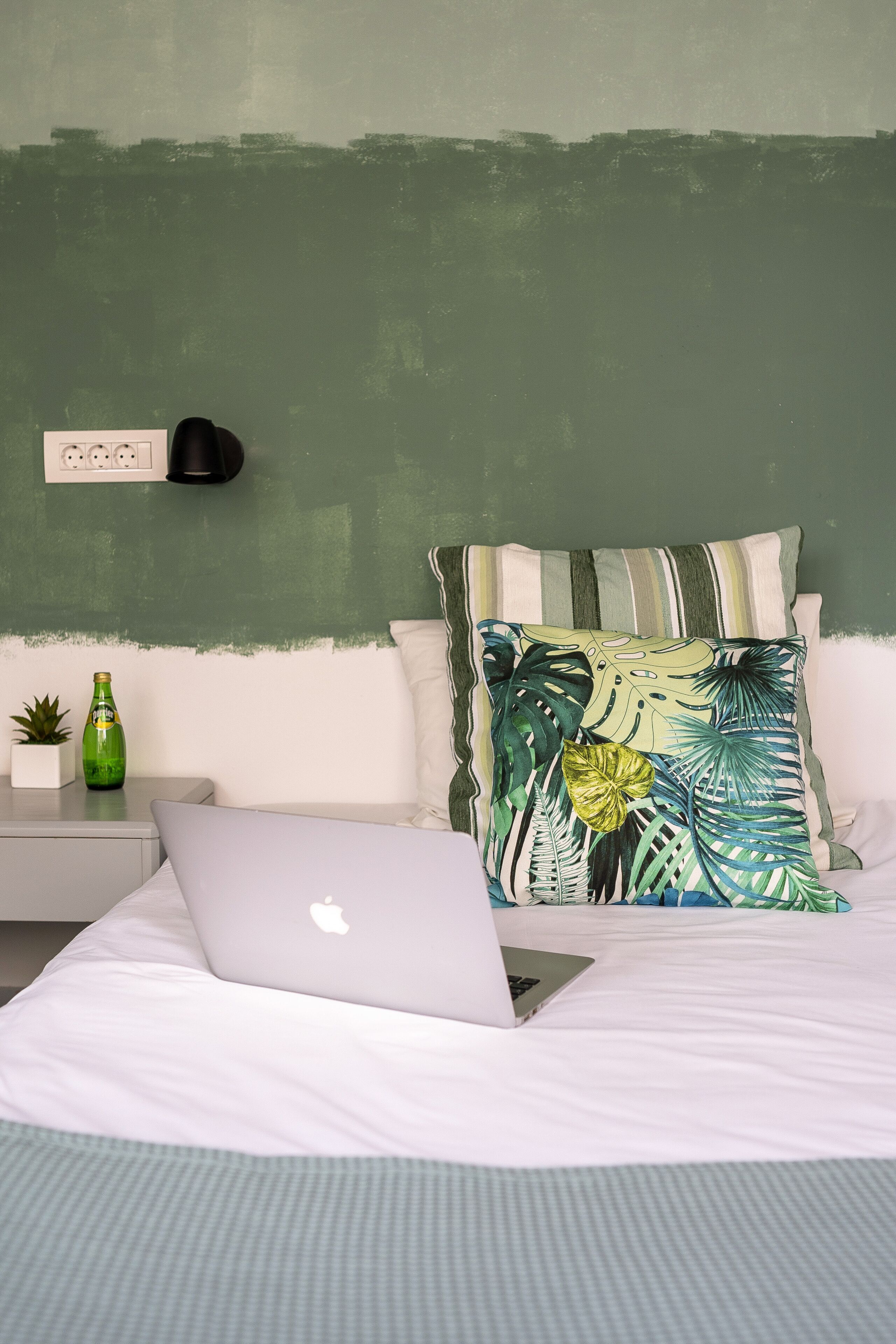 A neatly arranged bedroom with a bed, a laptop, a green bottle, and a small plant on a bedside table.