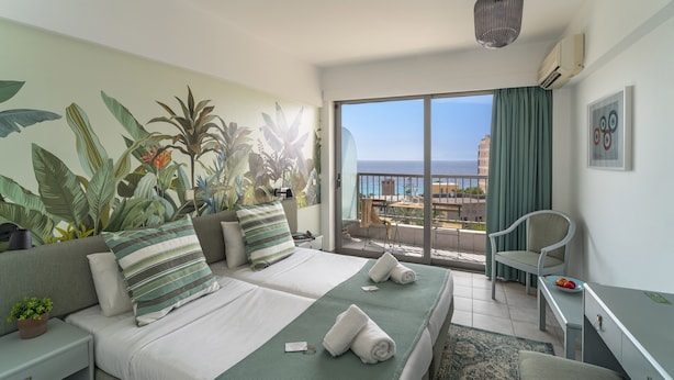 A hotel room with two beds, a balcony with a view of the sea, and a dining area with a table and chairs.