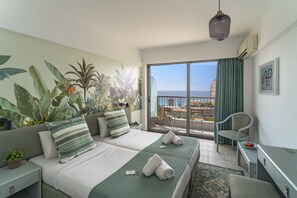 A hotel room with two beds, a balcony with a view of the sea, and a dining area with a table and chairs.