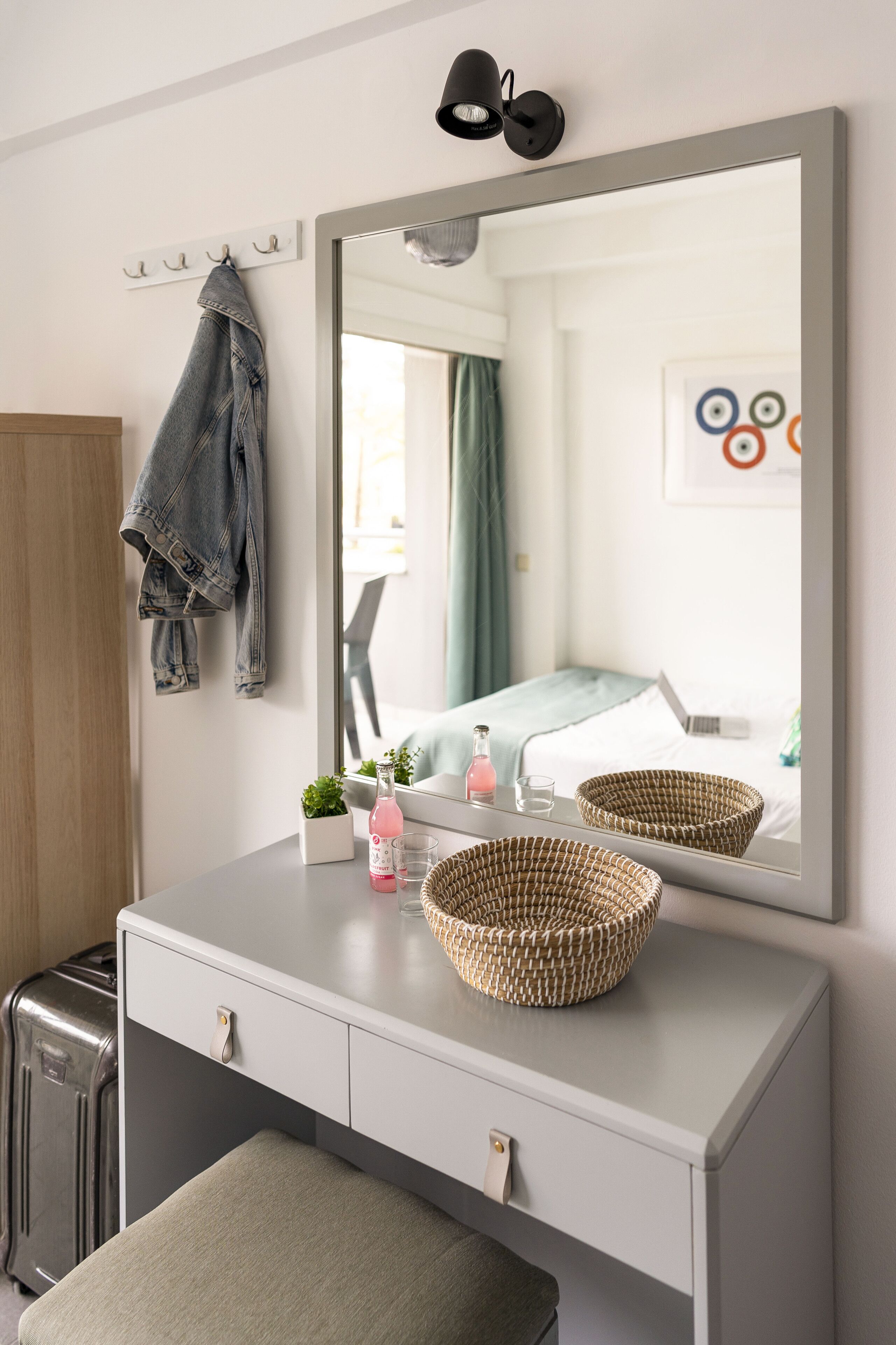 A hotel room with a large mirror, a bedside table with a lamp, a suitcase, and a bench.