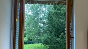 A window with open shutters, revealing a view of a green garden and trees.