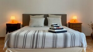A neatly made bed with striped bedding, two pillows, and a stack of folded towels on the headboard. There are two bedside tables with lamps, and a vase with dried flowers on the floor beside the bed.
