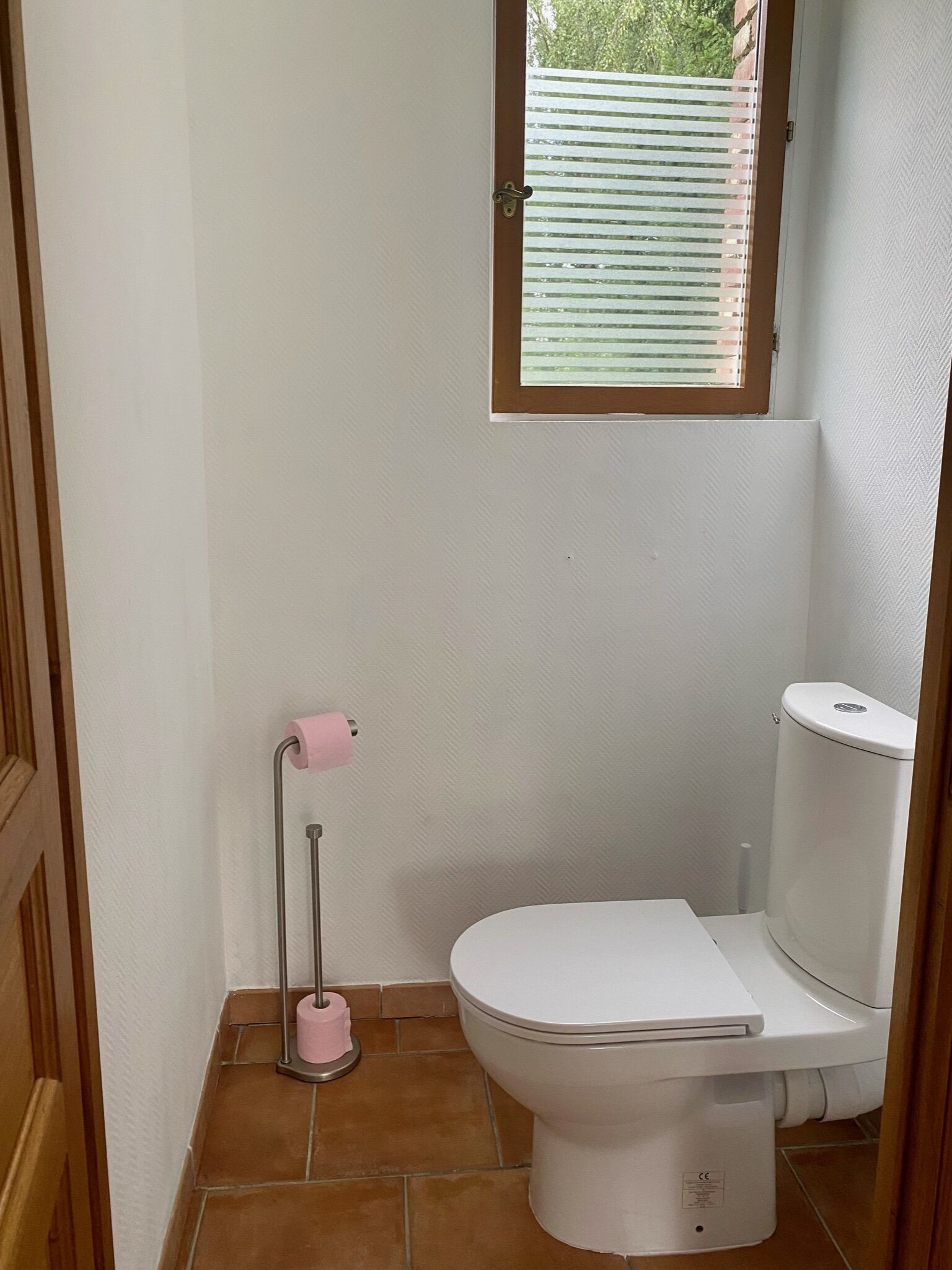 Een compacte, schone badkamer met toilet, een roze toiletrolhouder en een raam met zonwering.