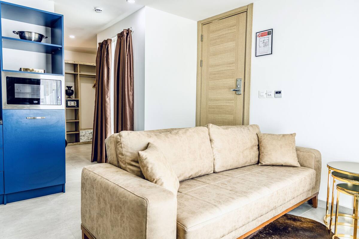 Un salon avec un canapé beige, un meuble bleu contenant des appareils de cuisine, une porte en bois et une petite table ronde.
