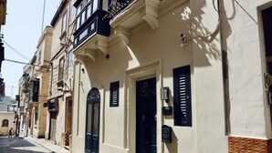 A street view of a building with a beige facade, black shutters, and a balcony with a wrought iron railing. - The Mike Hotel (Cospicua)
