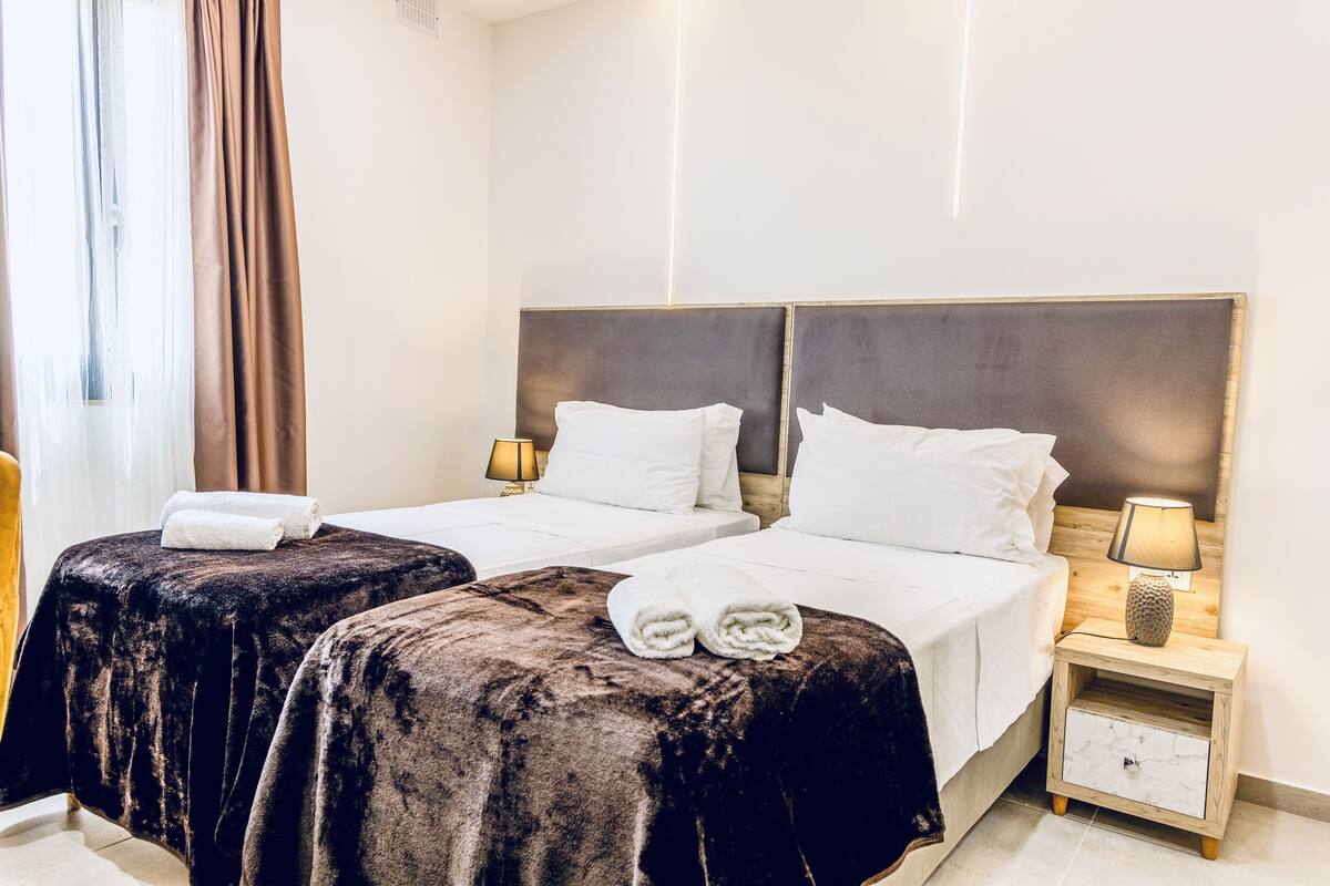 Une chambre d’hôtel avec deux lits, chacun doté d’une tête de lit sombre et de draps blancs. On y trouve une table de chevet avec une lampe et un petit tiroir.