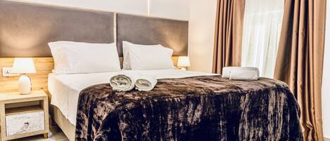 Una habitación de hotel con una cama, dos mesitas de noche, un cabecero y una ventana con cortinas.