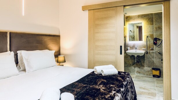 A hotel room with a bed, a nightstand, a lamp, a robe, and a bathroom visible through an open door. - The Mike Hotel (Cospicua)