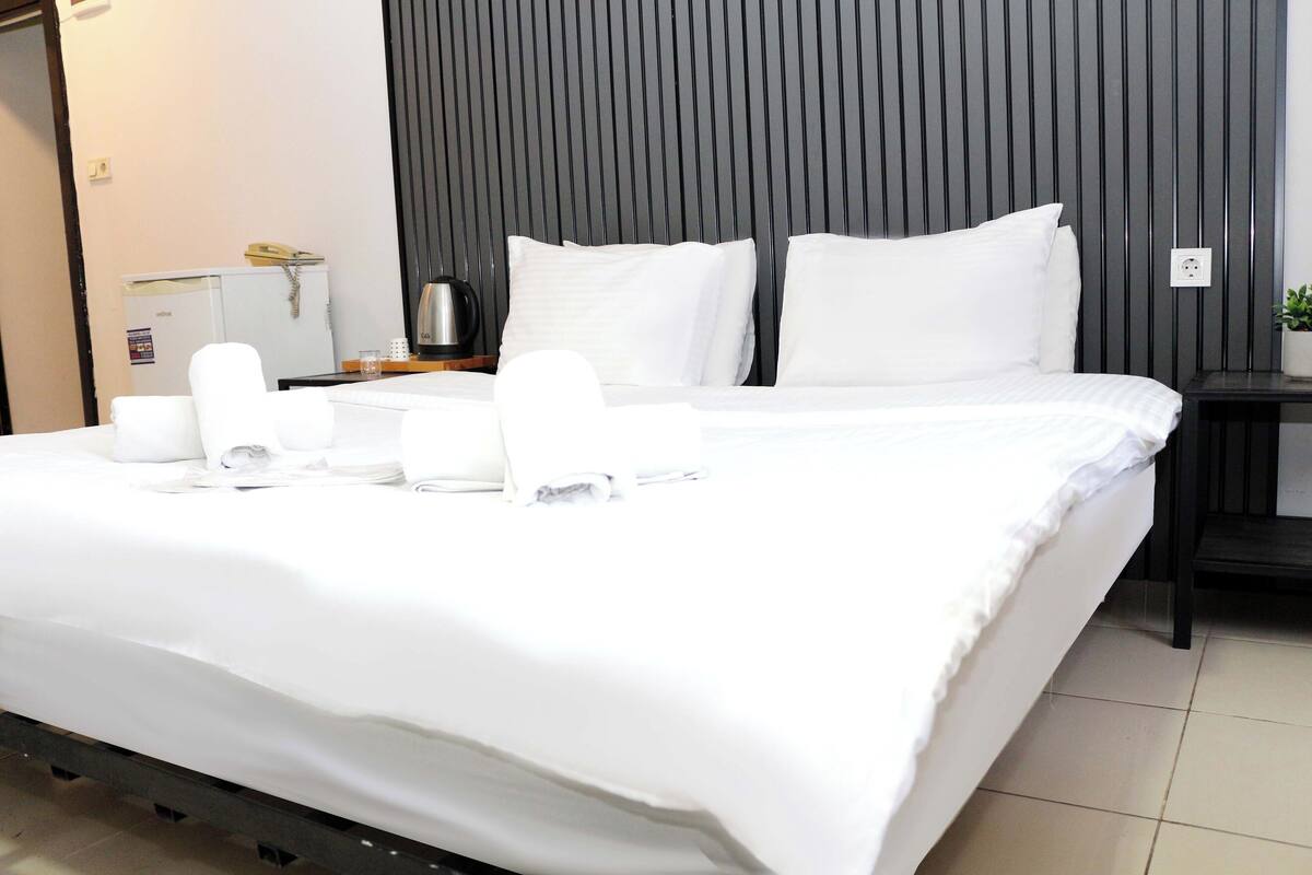 A neatly made bed with white linens and pillows, a small bedside table with a kettle, and a wall with vertical wooden slats.