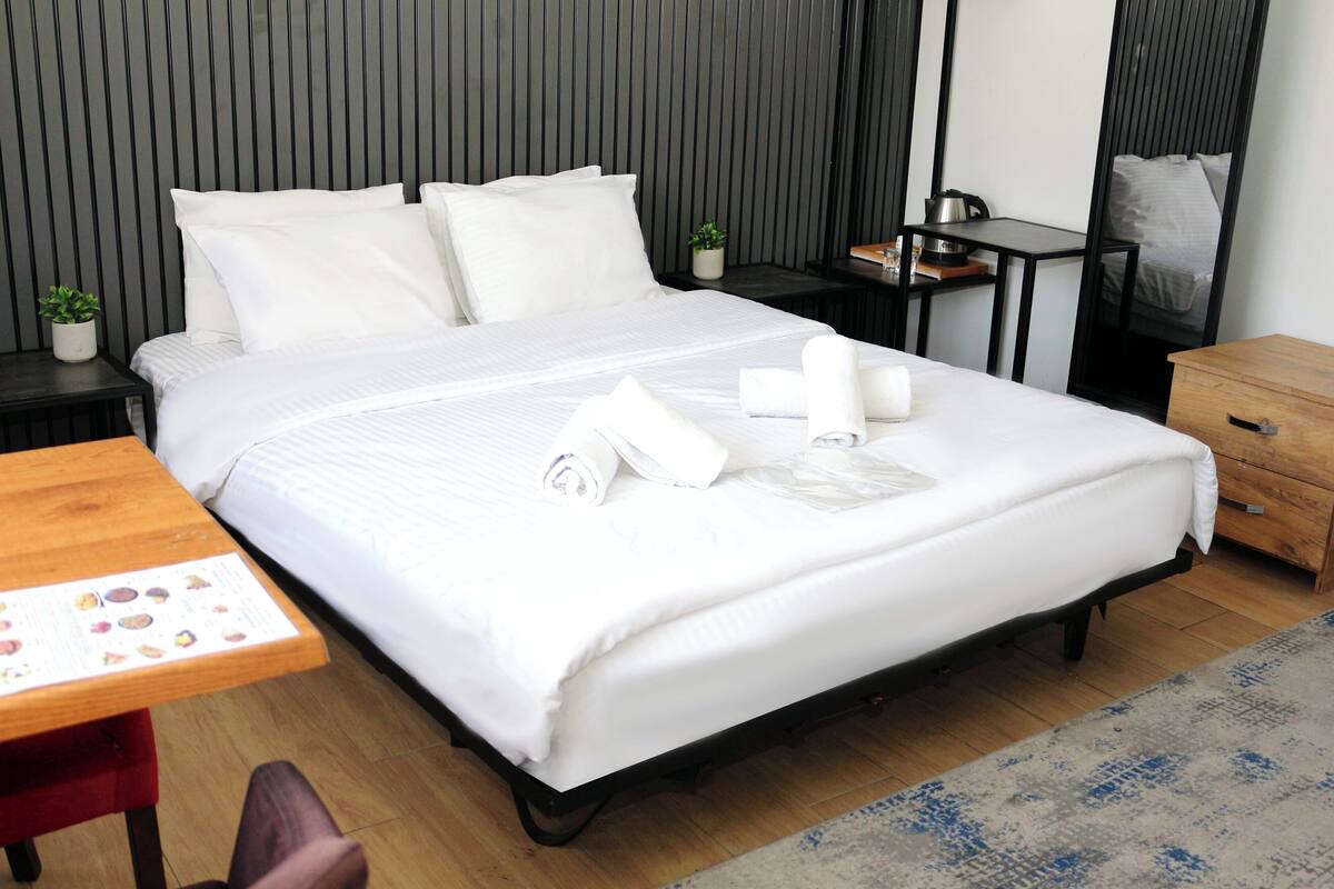 A neatly made bed with white linens and towels, a small wooden table with a menu, and a wooden nightstand with a plant.