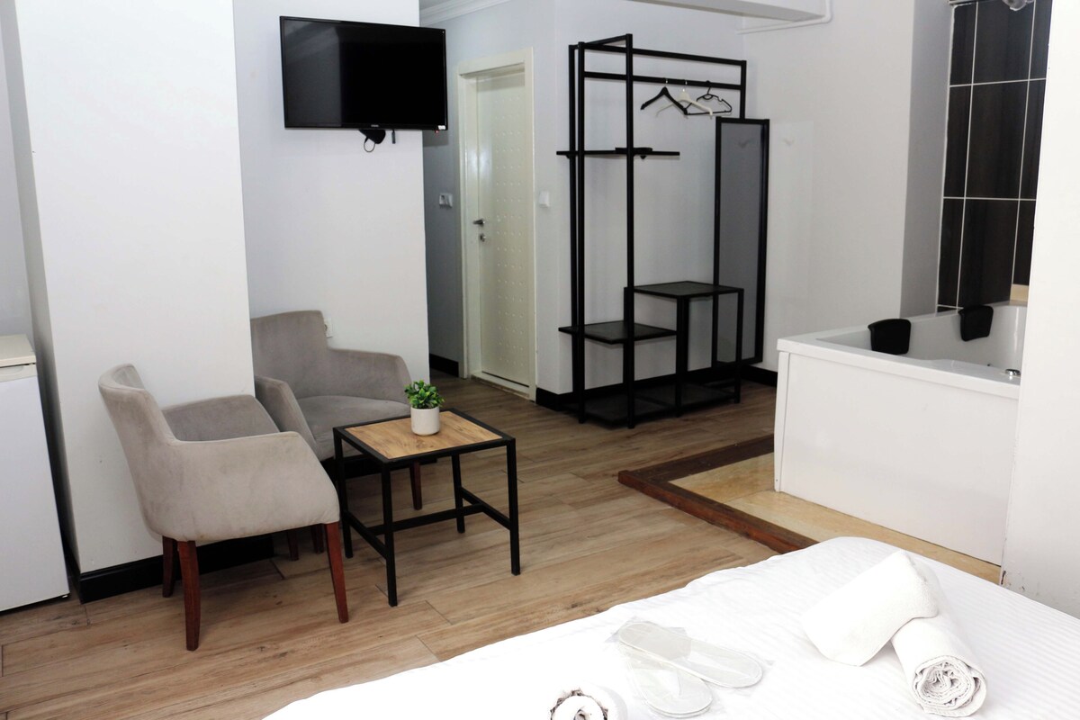 Ein Hotelzimmer mit einem Bett, einer Sitzecke mit Sessel und Tisch, einem an der Wand befestigten Fernseher, einem schwarzen Metallschrank und einer weißen Badewanne.