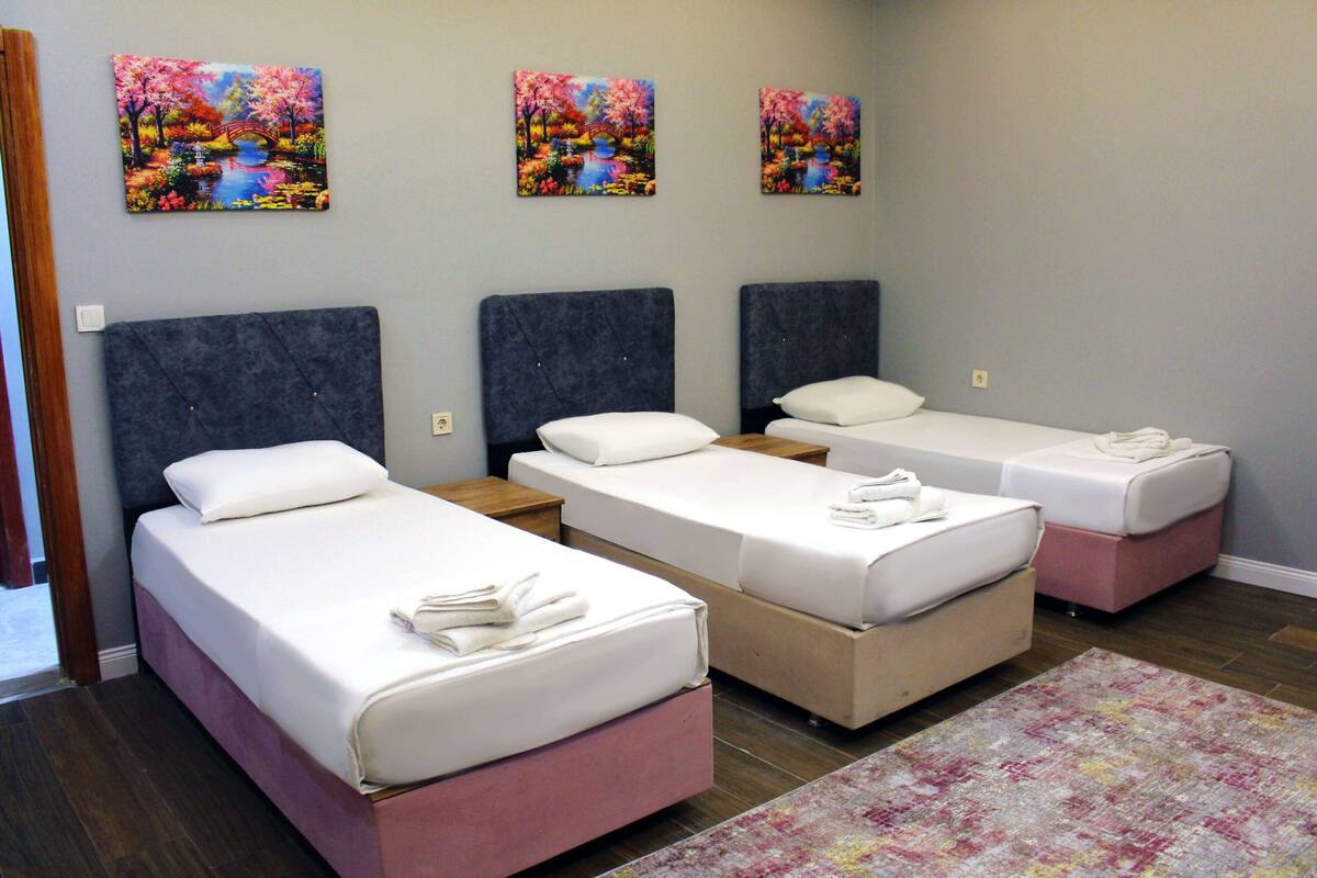A hotel room with three single beds, a wooden nightstand, a pink ottoman, and a colorful rug.