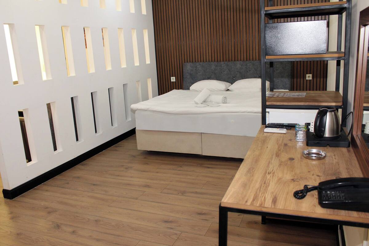 A hotel room with a bed, wooden floor, a desk with a telephone, a kettle, and a shelf with a mirror.