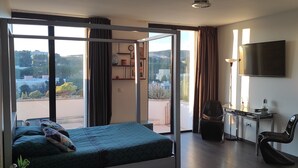 Ein Schlafzimmer mit Bett, Schreibtisch, Sessel und Fernseher, mit Blick ins Freie durch eine Schiebetür.
