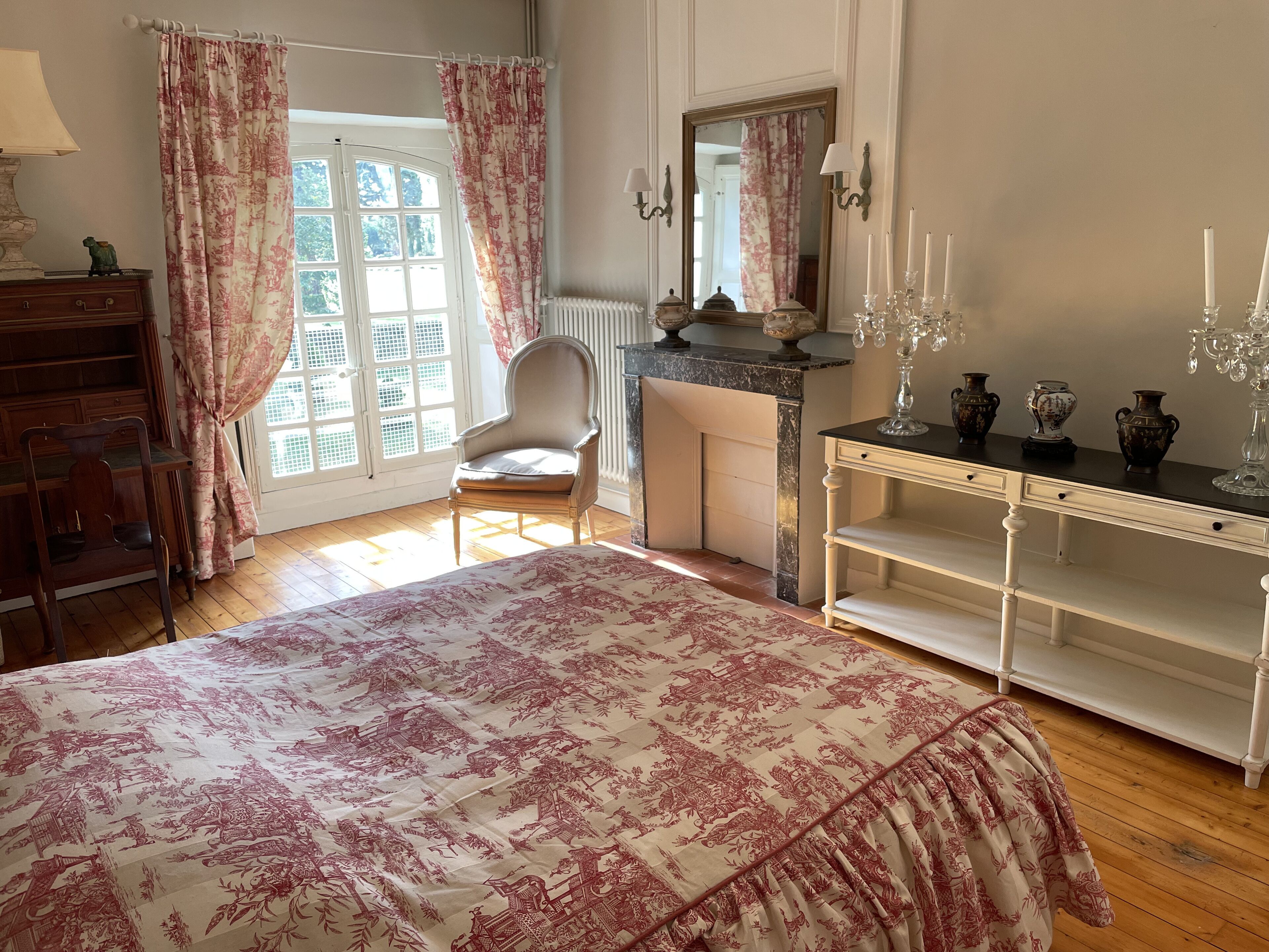 Un dormitorio con cama, una silla, un aparador, una tocador con espejo y una ventana con cortinas.