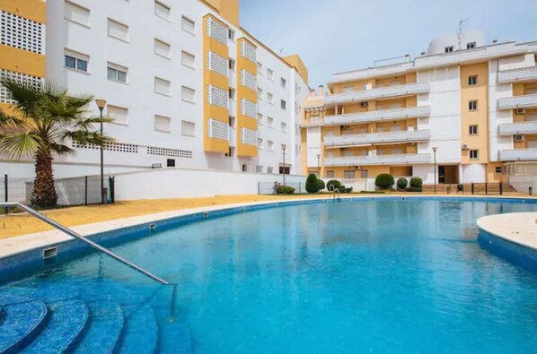 A swimming pool with a clear blue water surface, surrounded by a white building with balconies and a yellow facade.