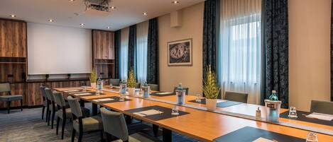 A conference room with a long table, chairs, a projector, and a large window with curtains.