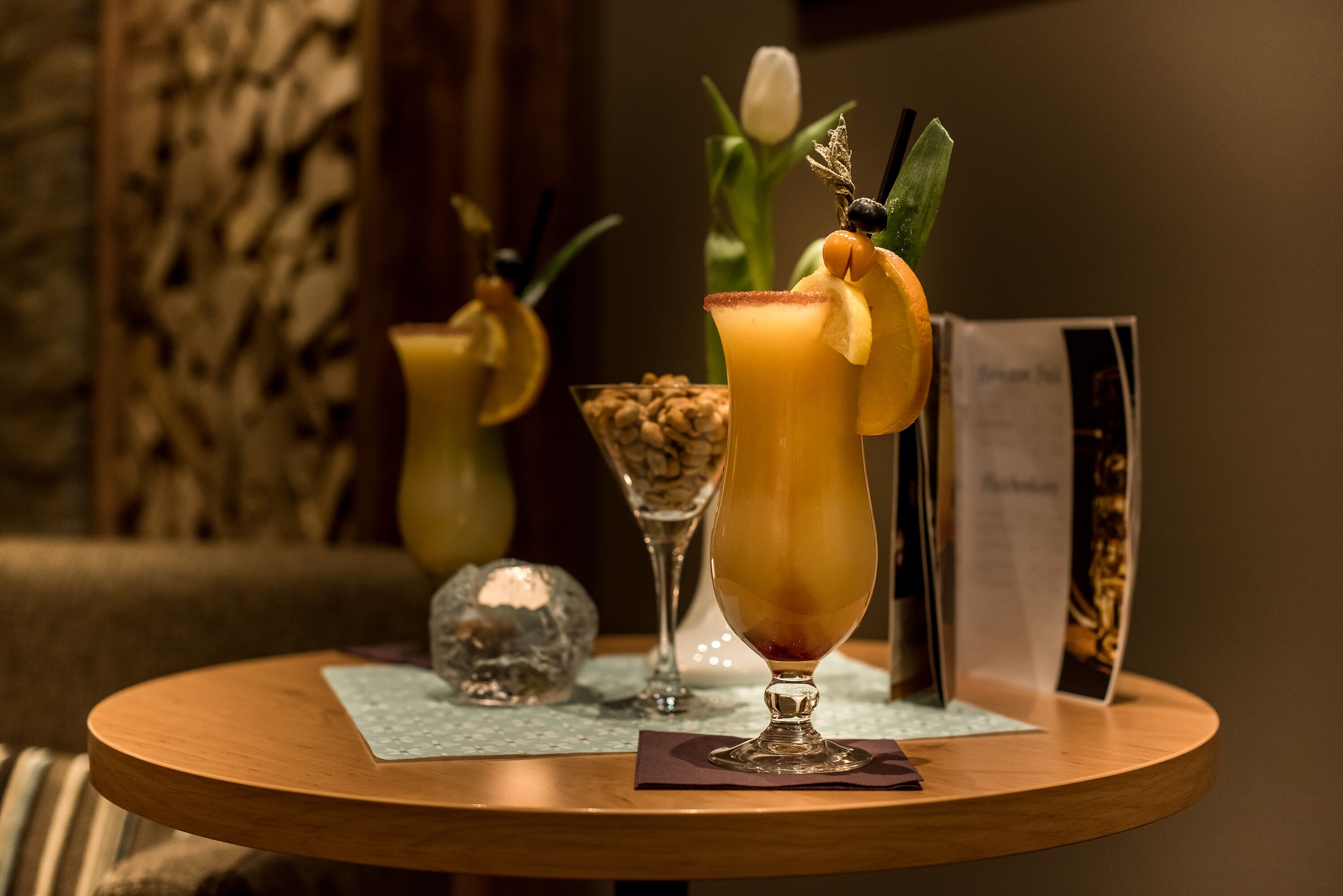 Une table ronde en bois, sur laquelle reposent deux cocktails décorés de fruits et un menu.