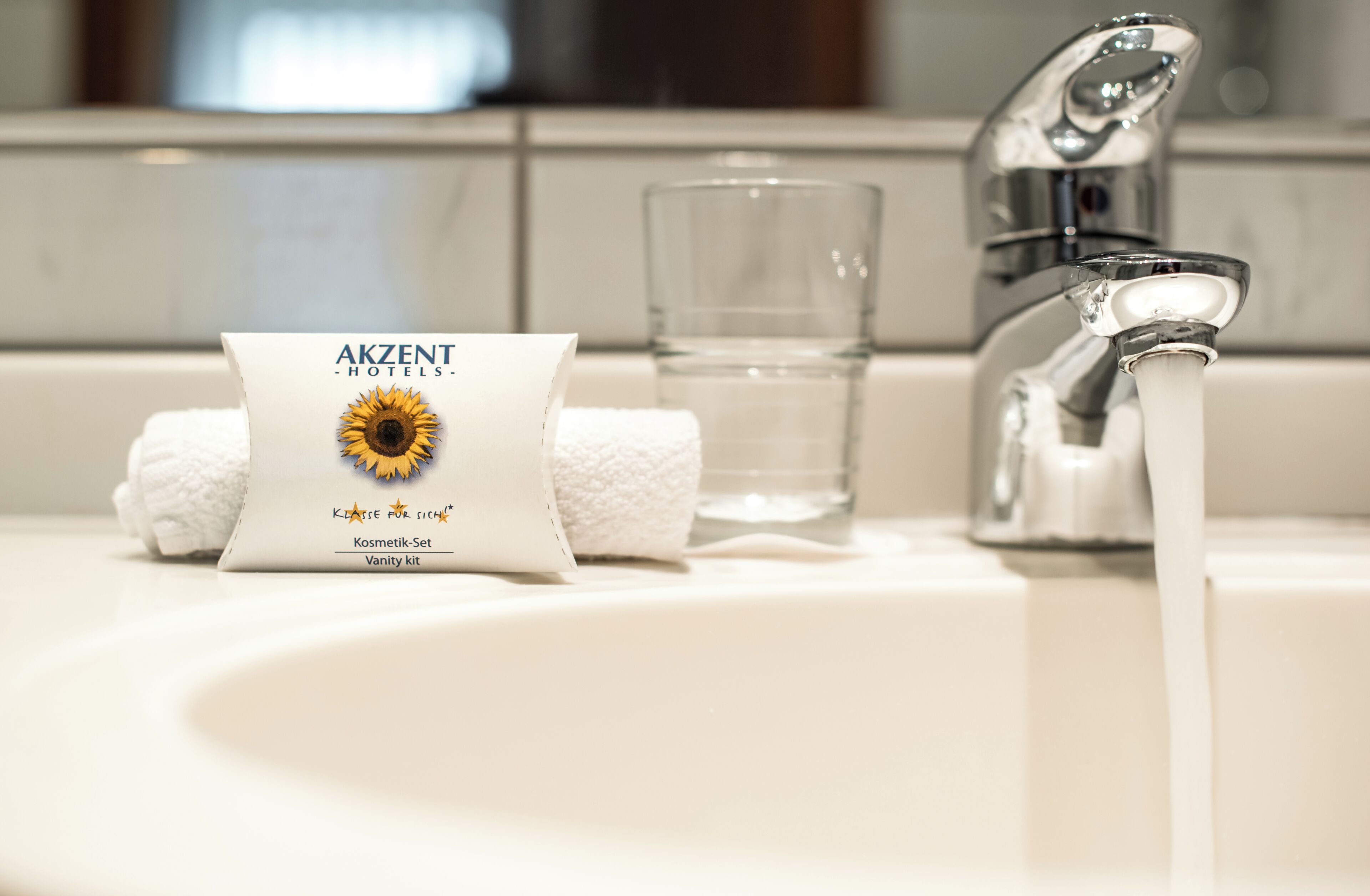 Un lavamanos de baño con una toalla, un vaso de agua y un paquete con la etiqueta 'AKZENT Hotels - Klassic für Sichei'.