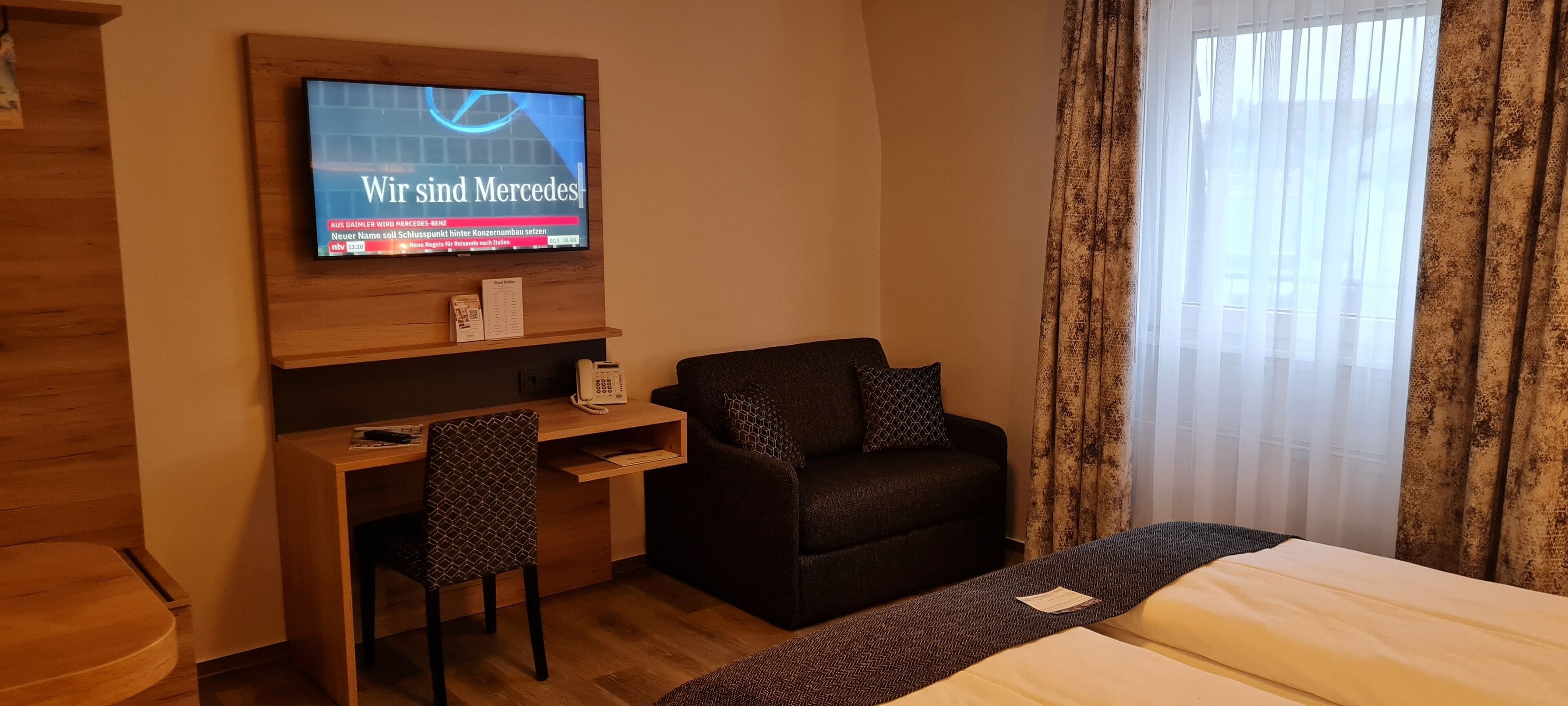 A hotel room with a bed, a desk with a chair, a television mounted on the wall, and a window with curtains.