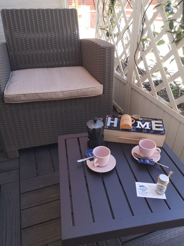 A wicker chair with a cushion, a small table with two pink cups and saucers, a spoon, a small bottle, and a card.