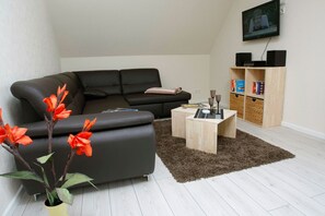 Uma sala de estar moderna com um sofá preto, uma mesa de centro, uma TV fixada na parede e uma estante com livros e objetos decorativos.