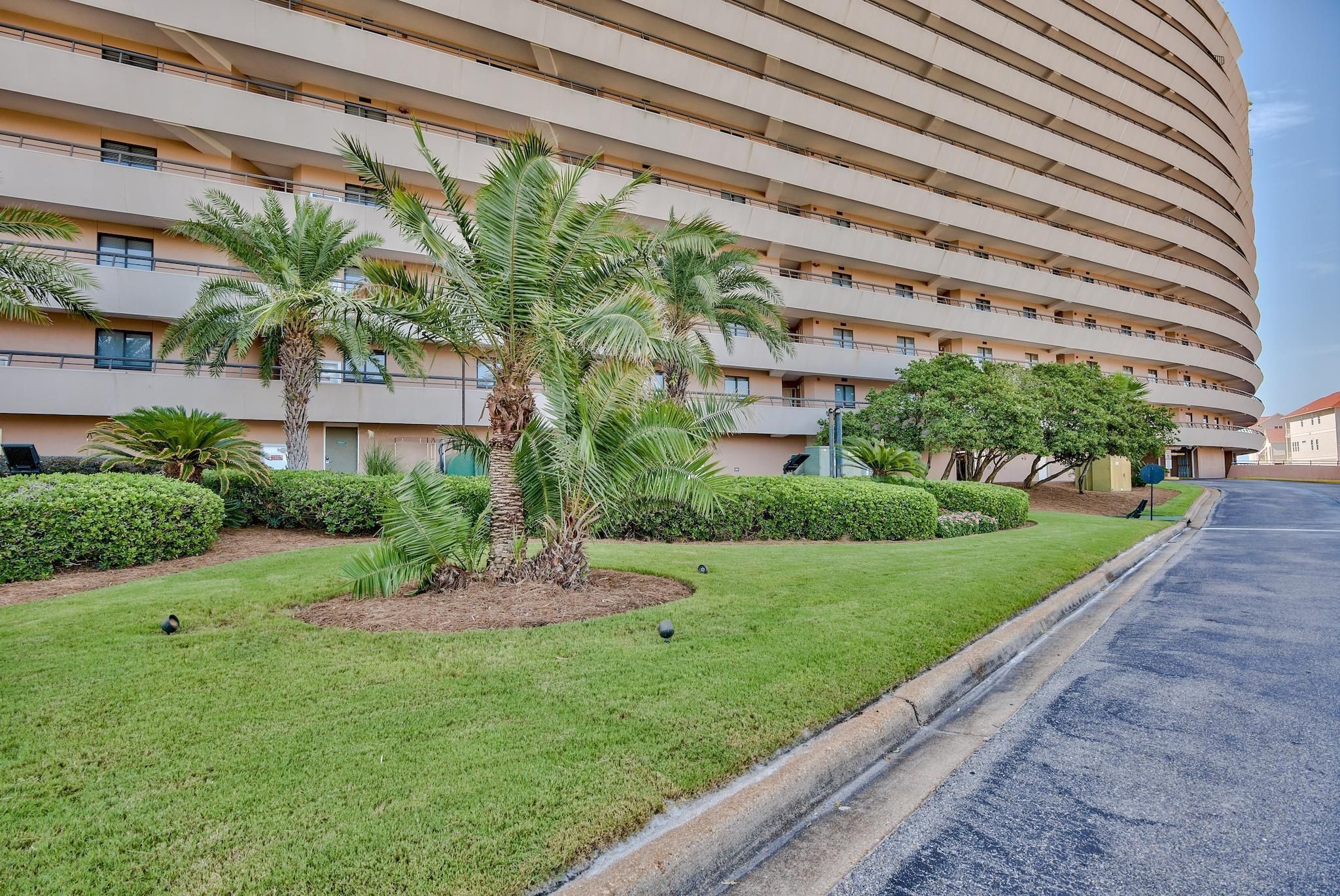 A modern hotel with a curved facade, multiple balconies, and well-maintained green lawns with palm trees.
