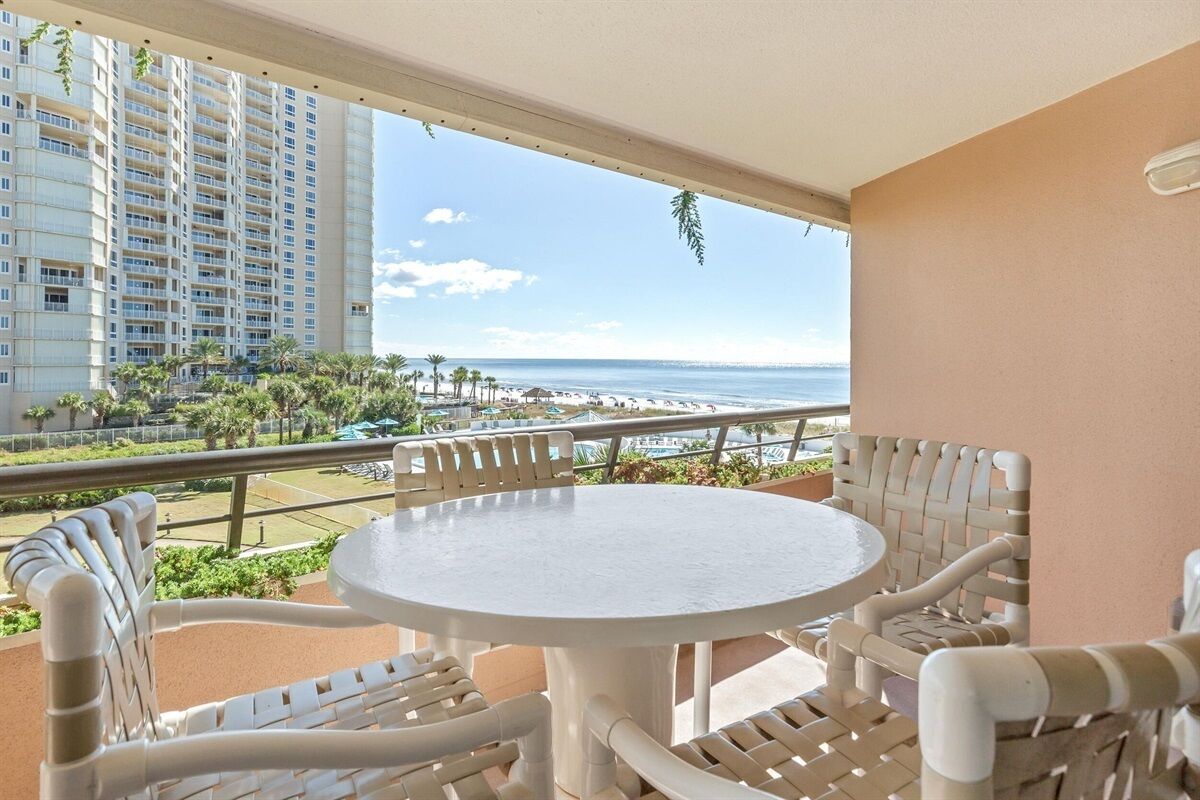 A balcony with a round table and white wicker chairs, overlooking a beach and high-rise buildings.