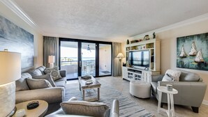 A modern living room with a sofa, armchairs, a coffee table, and a TV. There is a balcony with a view. - Edgewater 409 - Gulf Views! Beach & Pool Access! (Miramar Beach)
