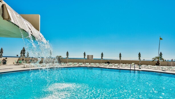 A swimming pool with water cascading from a structure, surrounded by lounge chairs and palm trees, with a clear sky in the background. - Edgewater 409 - Gulf Views! Beach & Pool Access! (Miramar Beach)
