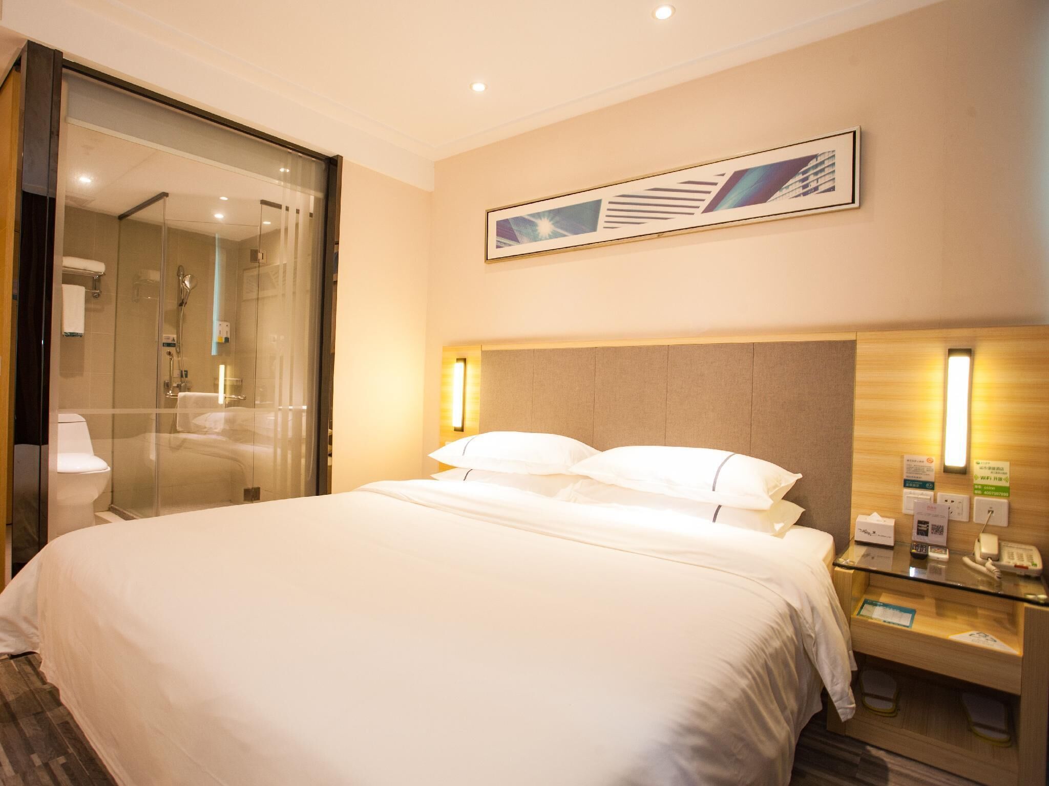 A hotel room with a large bed, a glass shower enclosure, a bedside table with a lamp, and a framed picture on the wall.