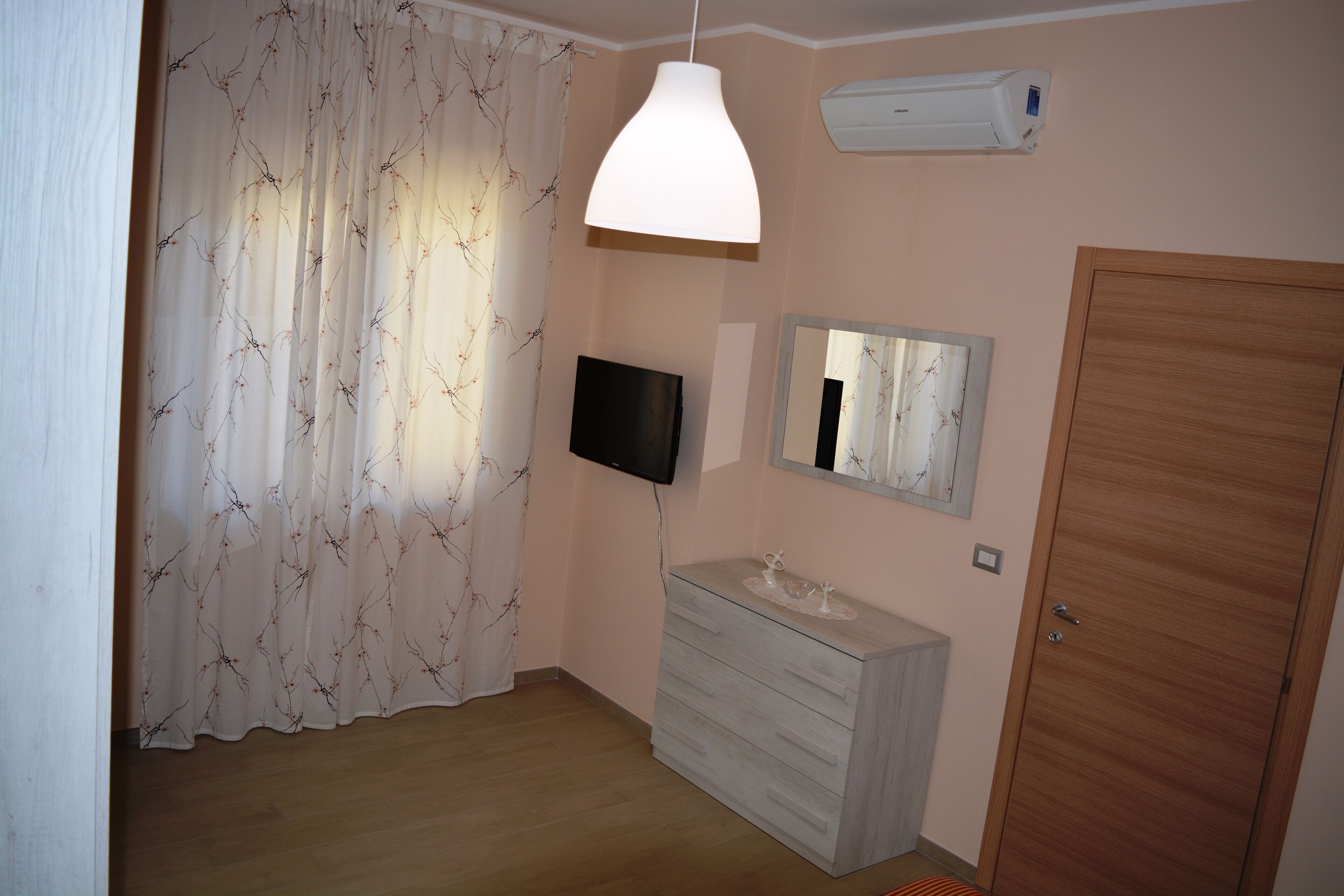 Une chambre avec un plancher en bois, une commode blanche, un téléviseur fixé au mur, un luminaire au plafond, une porte en bois et une fenêtre ornée de rideaux légers.