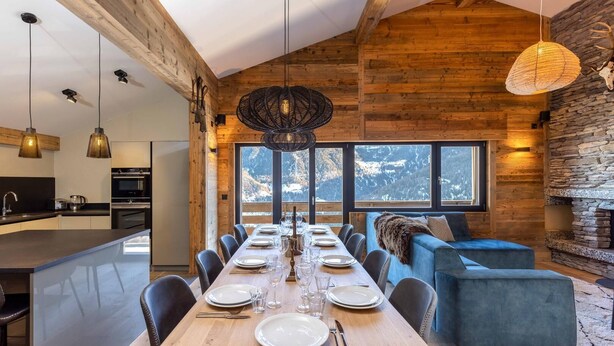 Un espace de repas moderne avec une longue table en bois, dressée pour six personnes, un canapé bleu et une cheminée en pierre.