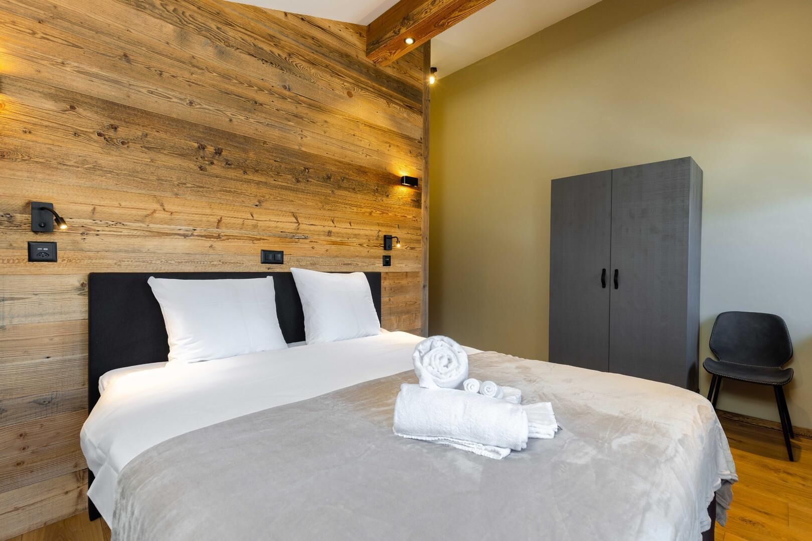 A neatly made bed with white linens and a grey blanket, a wooden headboard, a grey wardrobe, and a black chair.